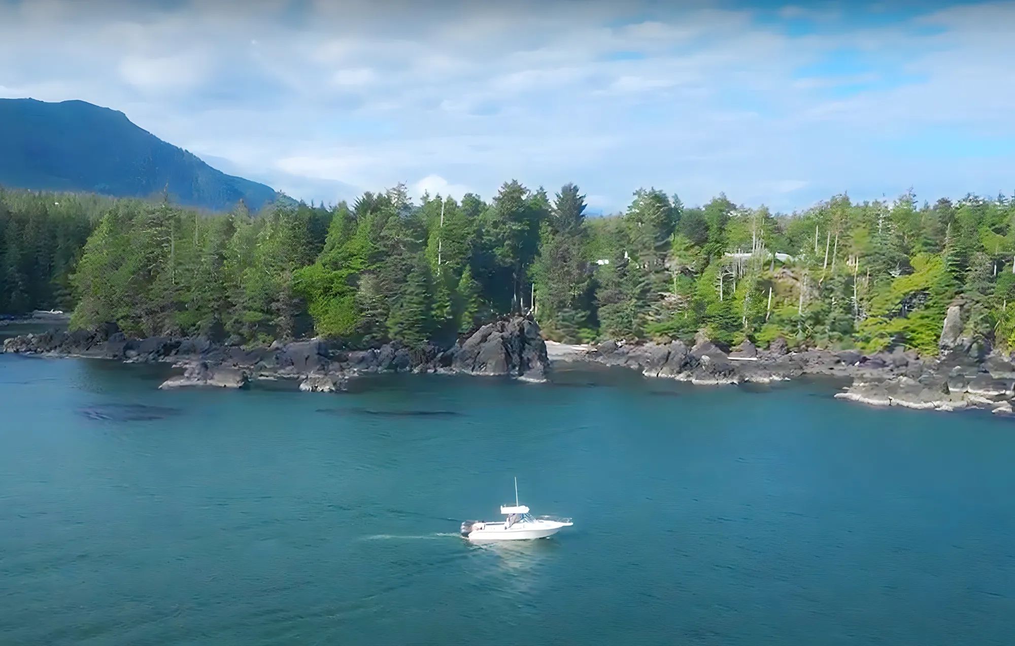 Boat on clear blue water near rocky forested coastline under a cloudy sky.