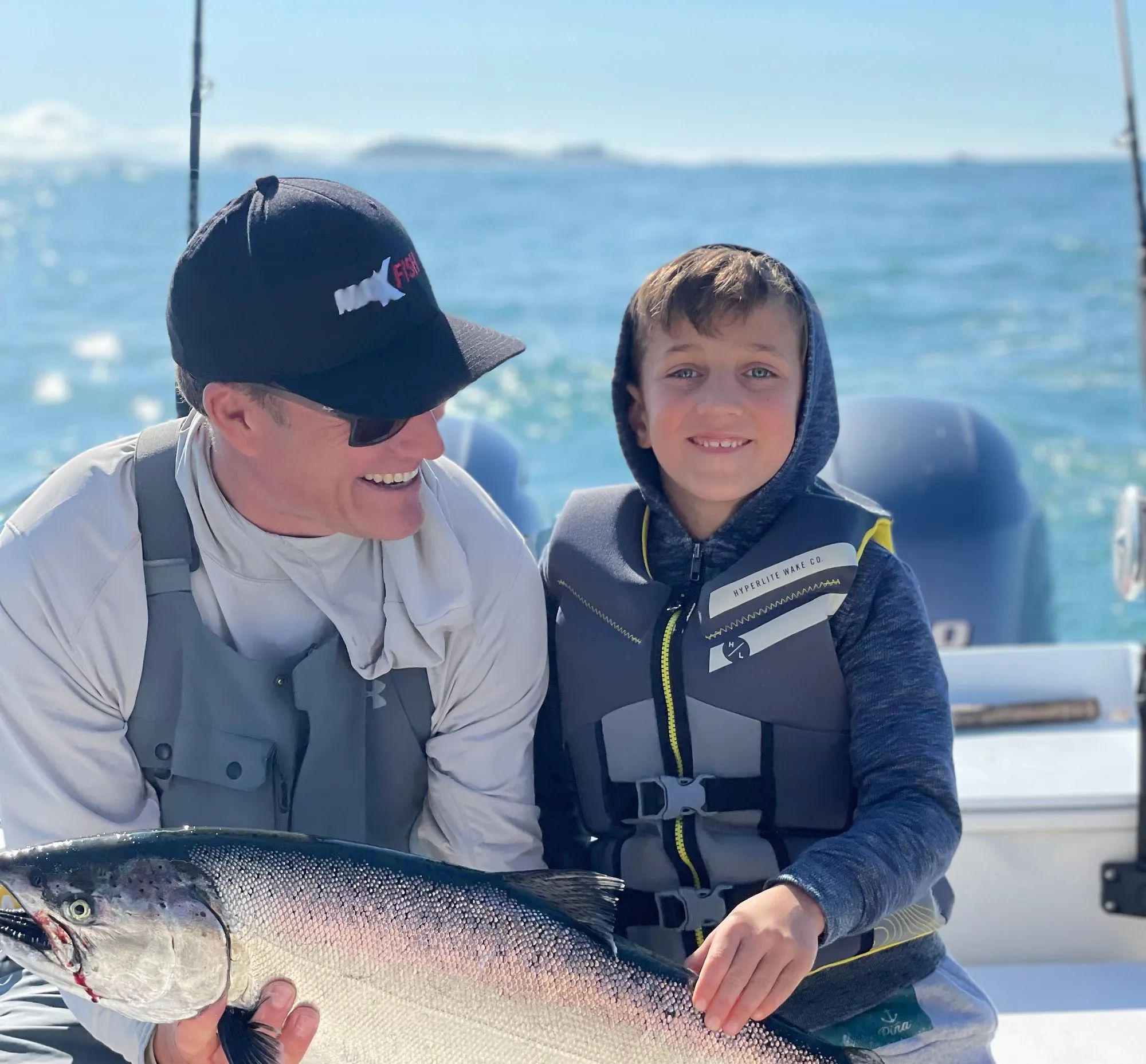 Father and son on a boat, holding a large fish, both smiling, wearing life jackets and caps, ocean backdrop.
