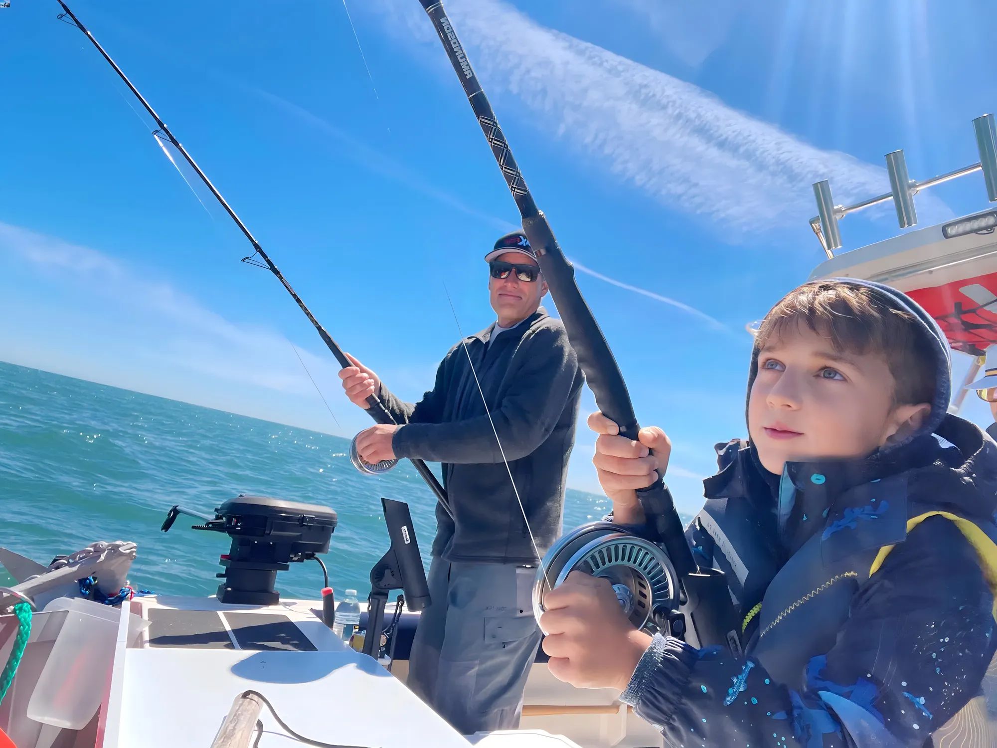 A man and child fishing on a boat under a clear blue sky, holding fishing rods, ocean in the background.