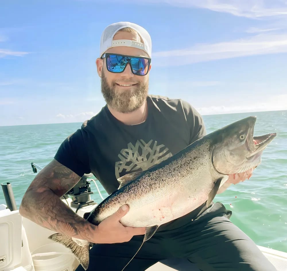 Person with beard and sunglasses holding a large fish on a boat in sunny ocean setting.