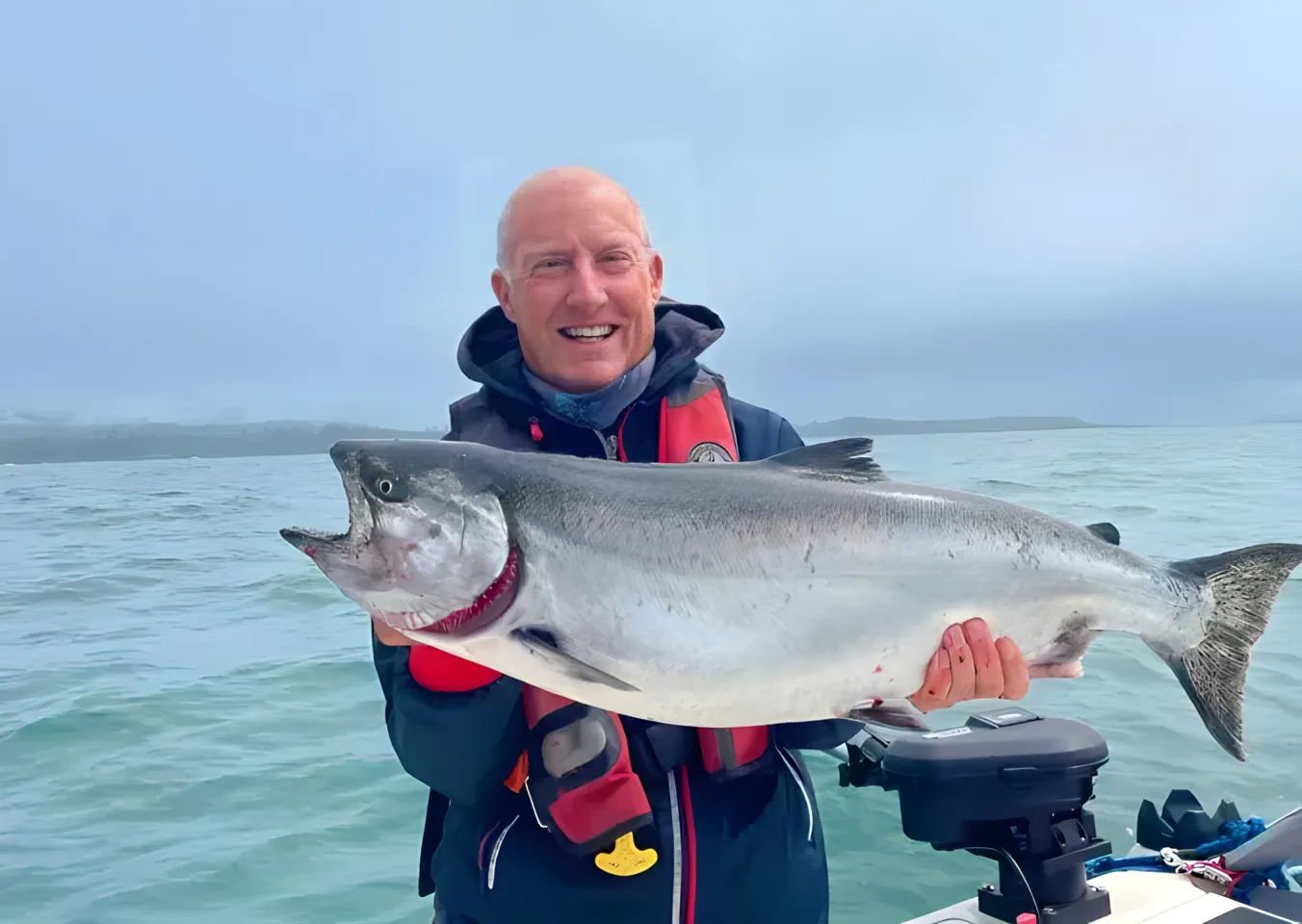 Person holding a large salmon on a boat, wearing a life jacket, with ocean and cloudy sky in the background.