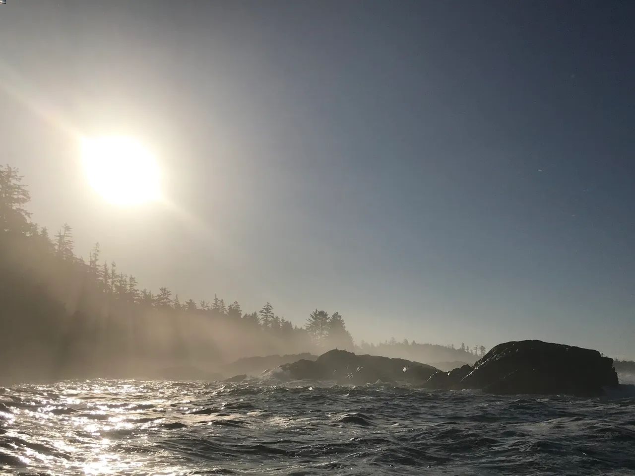 Sunlit ocean waves with rocky coastline and misty forest silhouette under clear sky.