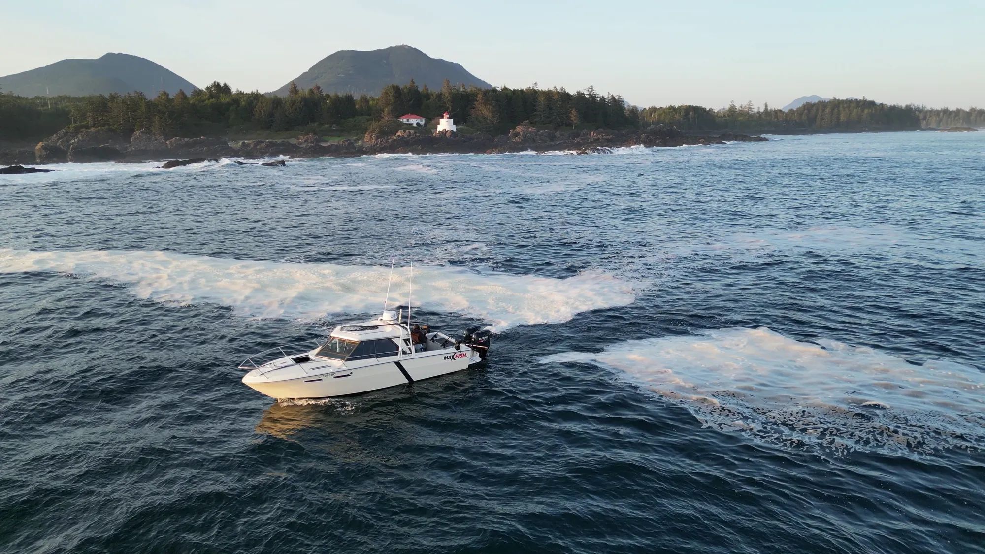 Boat on ocean near rocky shore, lighthouse, forest, mountains in background.