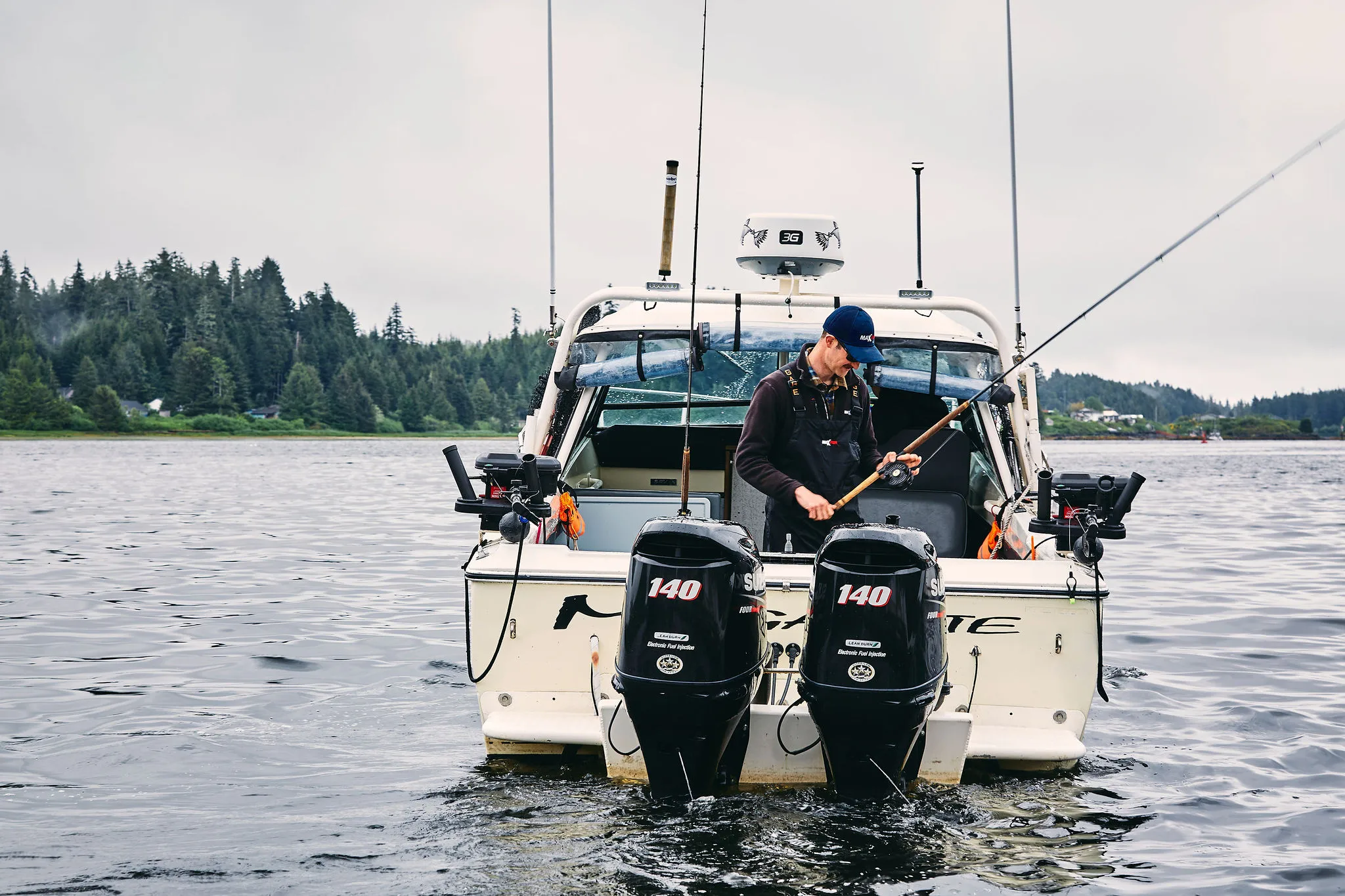 Fishing boat on a lake with a person preparing gear, surrounded by trees, overcast sky, dual outboard motors, calm water.