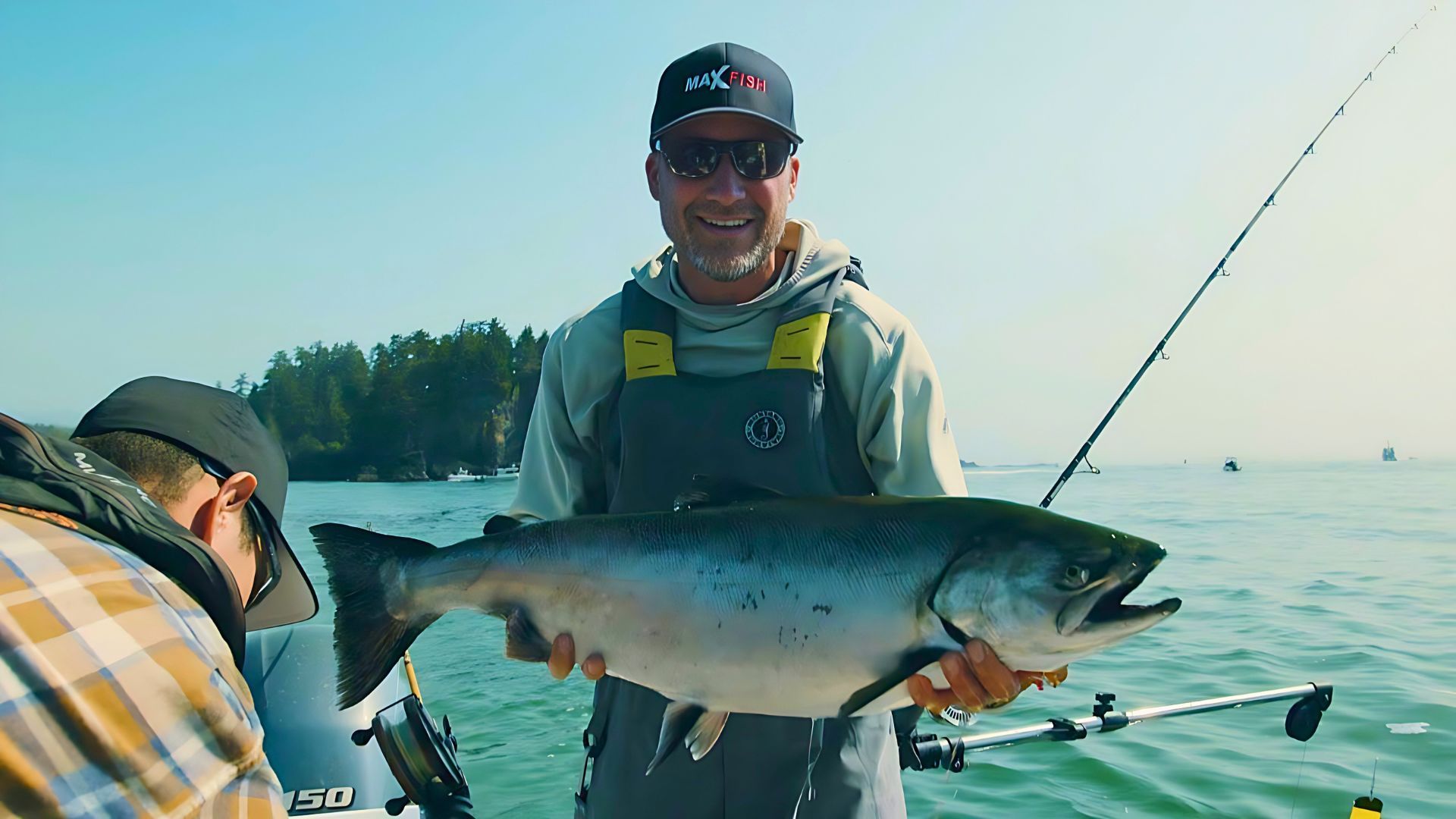 Person holding a large fish on a boat, wearing a cap and sunglasses. Fishing rod visible, with water and trees in the background.